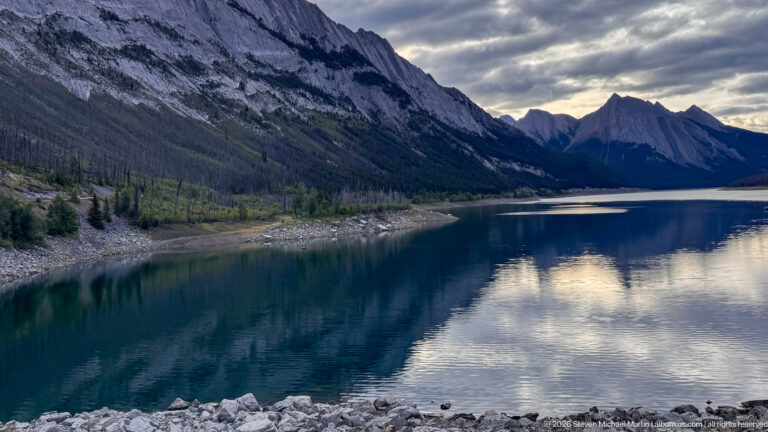 Medicine Lake narrowing between steep gray mountains with exposed gravel shoreline and dark reflective water.