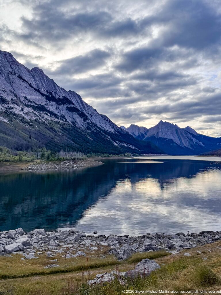 Medicine Lake beneath layered clouds with broad exposed rocky shoreline and steep mountain walls reflected in dark water.