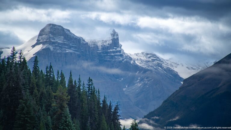 A steep layered mountain rises above conifers with snow and low cloud clinging to its upper slopes