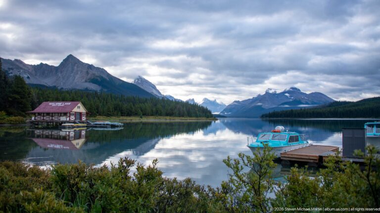 The red-roofed Maligne Lake Boathouse, docks, and boats sit on reflective water beneath mountain ridges