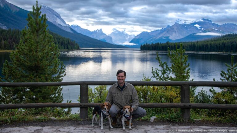 A seated visitor poses with two beagles before a railing overlooking Maligne Lake and mountains