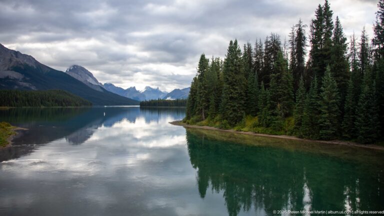 A conifer-covered peninsula projects into calm mountain water with strong reflections