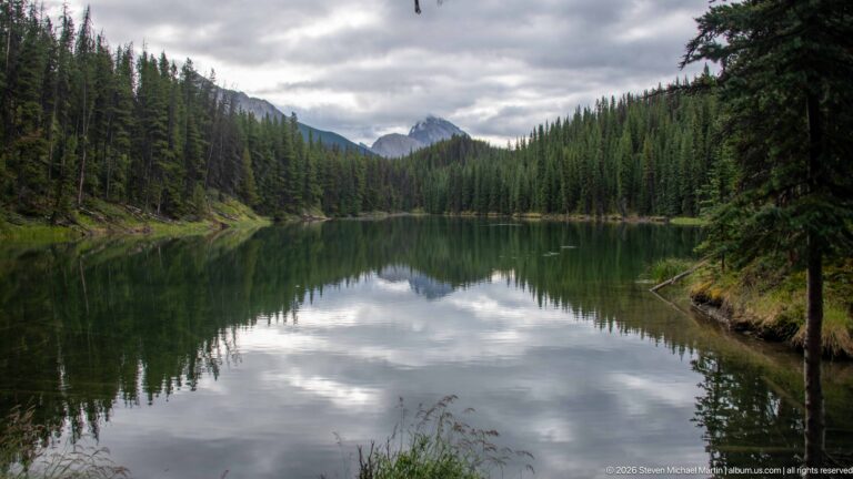 Still water reflects conifer forest and cloudy sky in a small enclosed mountain lake