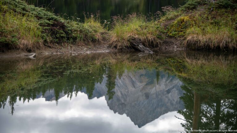 Still forest pond reflecting a steep gray mountain peak, conifers, and cloudy sky.
