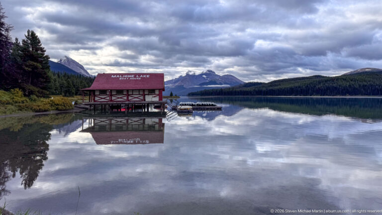 Red-roofed Maligne Lake Boat House and adjacent canoes reflected in calm water beneath cloudy mountain ridges.