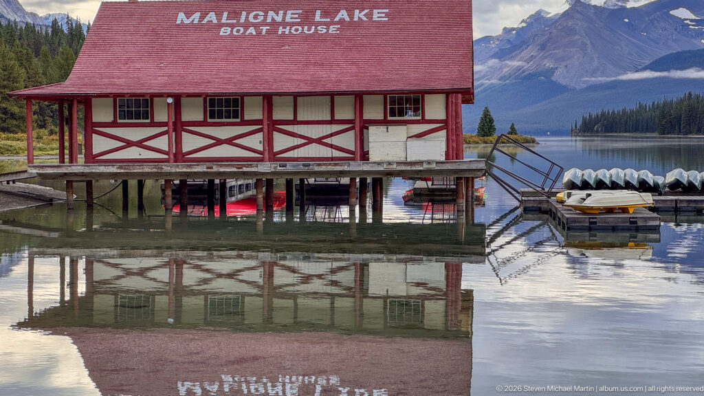 The timber-framed Maligne Lake Boat House fills the frame above its reflection, with dock structures and stacked canoes beside it
