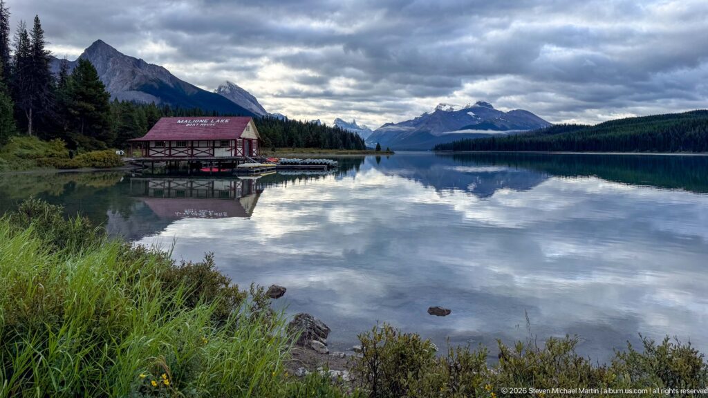The Maligne Lake Boat House sits small at the left edge of a calm reflective lake beneath dramatic cloud and mountain scenery