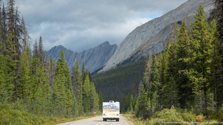 Vehicle traveling on a forest-lined mountain road beneath steep gray ridges in the Maligne Valley.