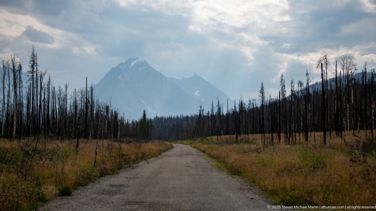 Straight paved road crossing burned forest toward a steep mountain beneath a partly cloudy sky.