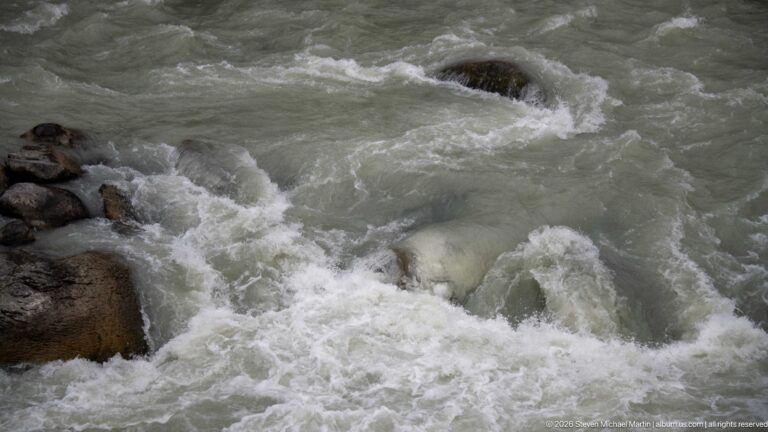 Close view of gray-green river water breaking into white foam around dark submerged boulders.