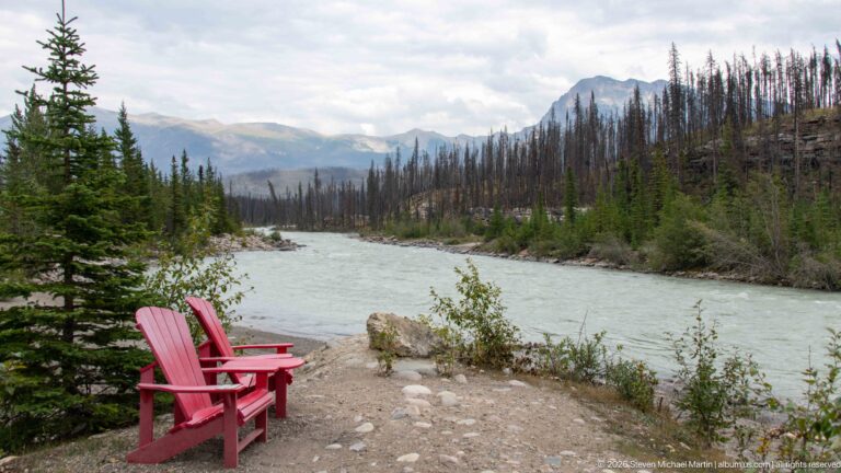 Two red Adirondack chairs facing a turquoise river, with conifers, burned forest, and mountains across the valley.