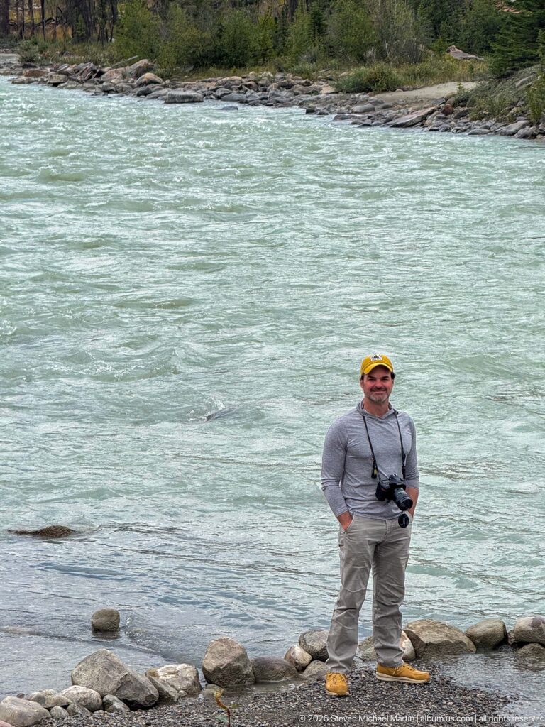 Visitor standing on rounded rocks at the edge of a broad pale turquoise mountain river.