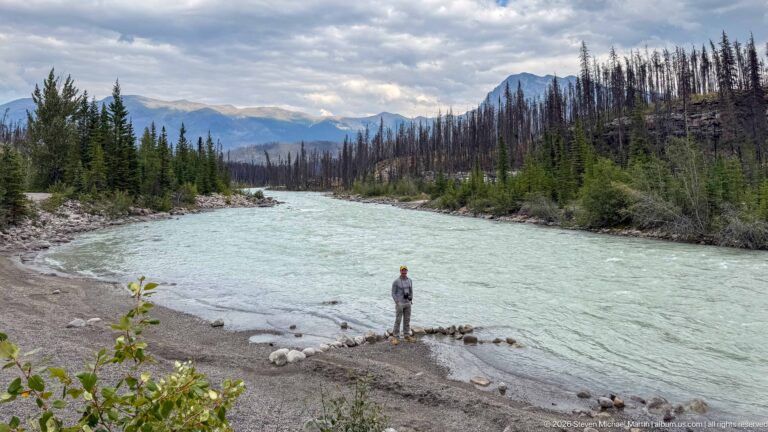 Wide turquoise river curving through a mountain valley, with a visitor and two beagles on the gravel shore.