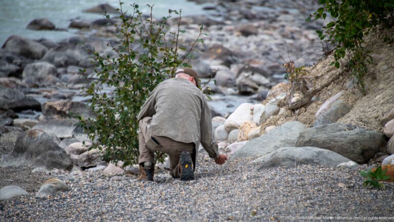 Visitor crouching on a gravel riverbank among large boulders, with flowing water visible beyond.