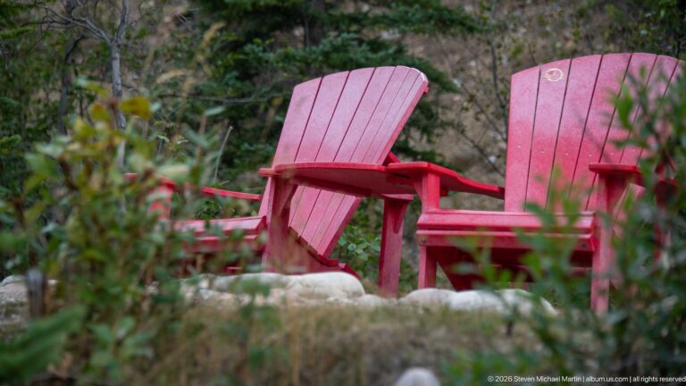 Two empty red Adirondack chairs among shrubs and conifer trees at a forested Jasper National Park viewpoint.