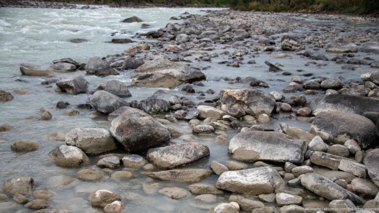 Pale turquoise river water flowing around rounded gray cobbles and boulders in a shallow rocky channel.