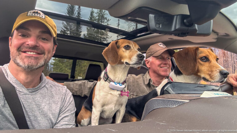 Two men and two beagles inside a vehicle, with conifer trees visible through the windows and glass roof.