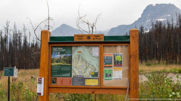 Wooden Fryatt Valley trailhead kiosk with maps and notices, backed by burned trees and steep mountains.