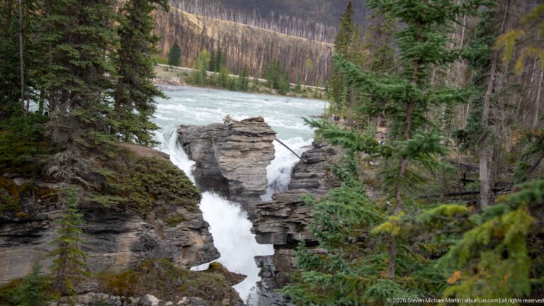 Turquoise river narrowing into a white cascade between layered rock outcrops, surrounded by evergreen forest.