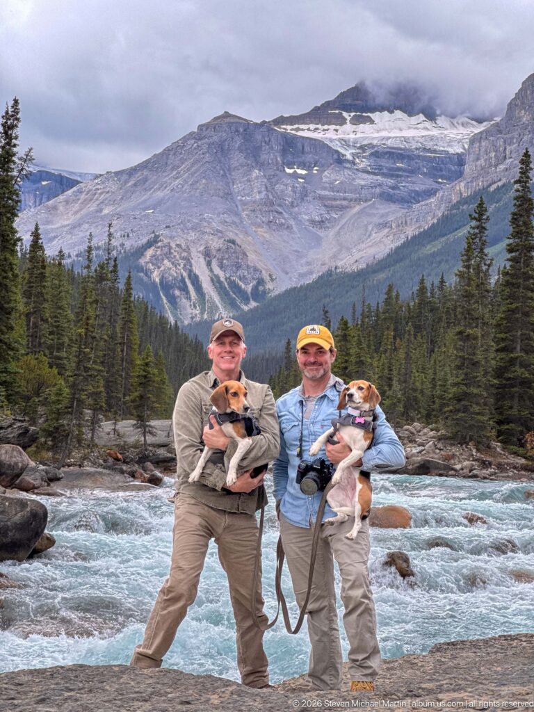 Two visitors with beagles standing beside rushing turquoise water beneath a broad gray mountain and low clouds.
