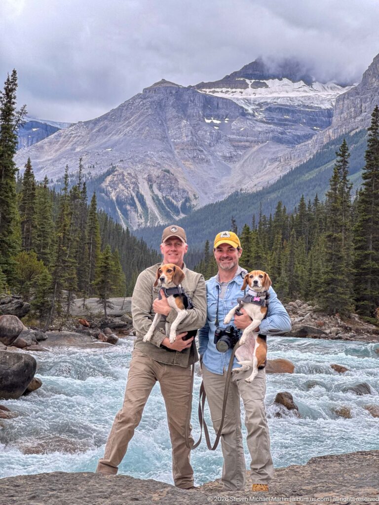 Two visitors holding beagles beside a turquoise mountain river, backed by conifer forest and a cloud-covered rocky slope.