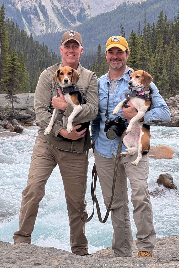 Two men holding beagles on a rocky bank beside fast turquoise water, with forested mountains behind them.