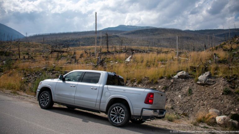 Silver pickup truck parked beside a road with extensive burned forest and mountain slopes under heavy clouds.