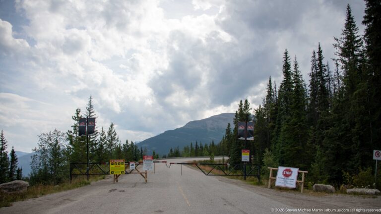 Barrier gate and warning signs closing a paved mountain road bordered by conifers beneath cloudy mountain slopes.