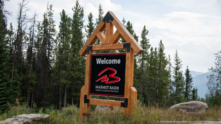 Timber-framed Marmot Basin welcome sign with red logo, surrounded by conifers and mountain scenery.