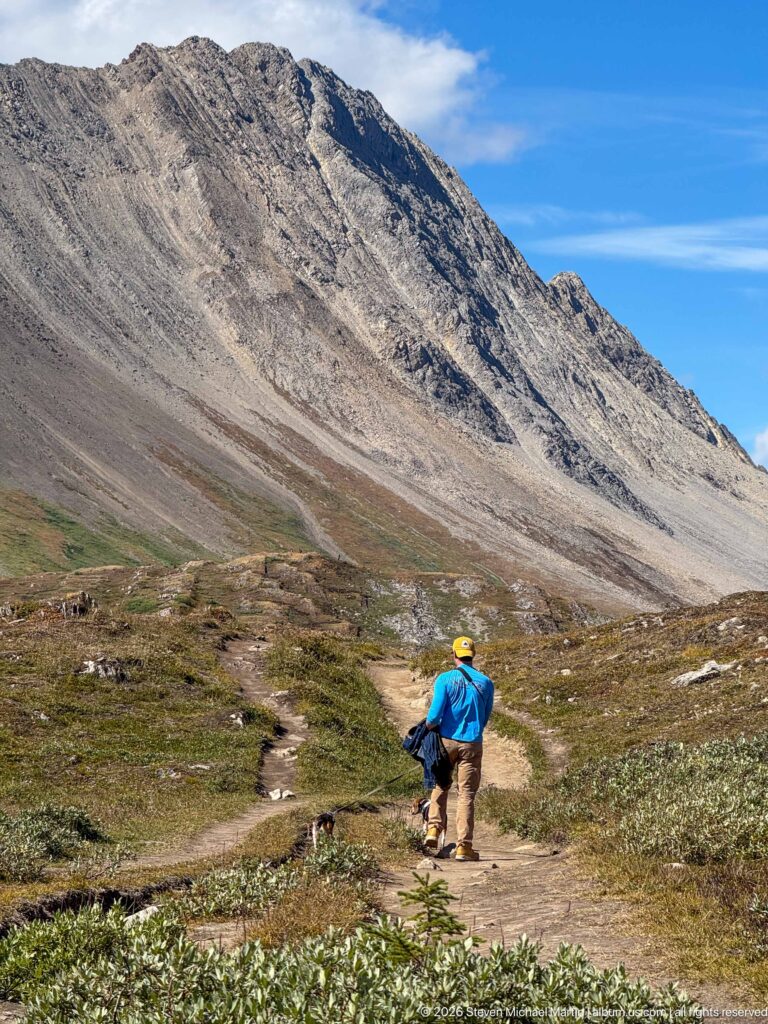 Hiker descending a dirt trail through alpine tundra beneath a broad gray scree-covered mountain slope.