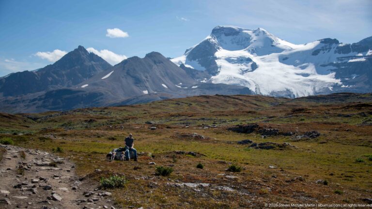 Small hiker and dogs beside an alpine trail beneath dark peaks and a large snow-covered mountain massif.