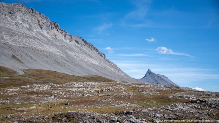 Rocky alpine pass below a steep pale-gray escarpment with a pointed mountain peak in the distance.
