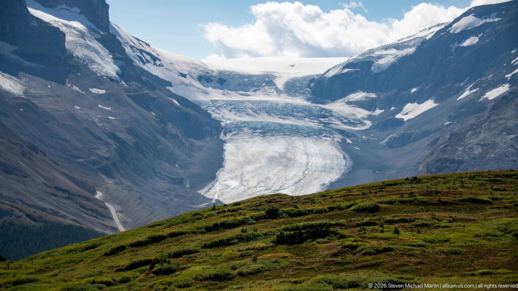 Large crevassed glacier descending between dark mountains above a rounded green alpine ridge.