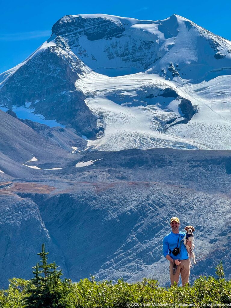 Hiker holding a beagle below a broad glacier and snow-covered mountain basin.