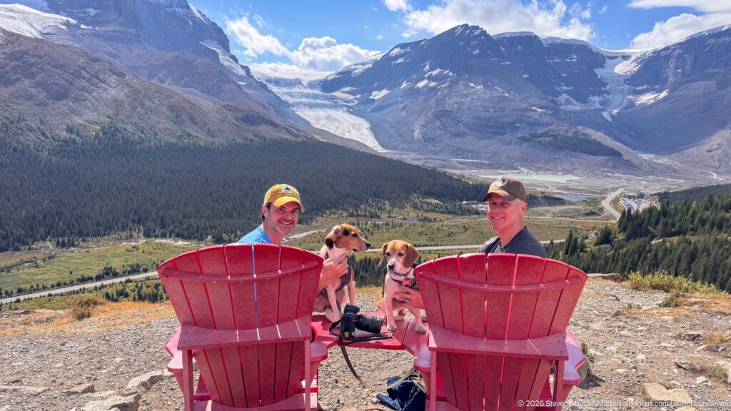 Two hikers with two beagles seated at red chairs above a glacier-filled mountain valley and roadway.