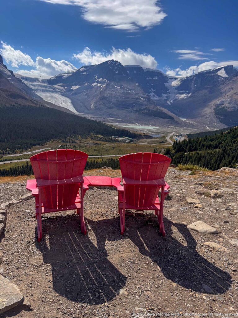 Two red Adirondack chairs facing a panoramic glaciated mountain valley from a rocky Wilcox Pass overlook.