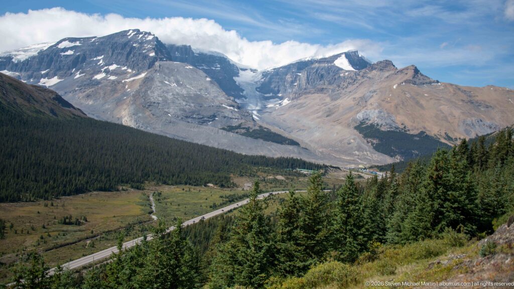 Icefields Parkway winding through a forested mountain valley below high gray peaks and patches of glacier ice.