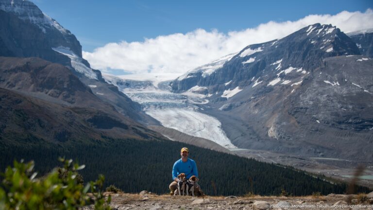 Hiker with two leashed beagles standing at an overlook with a large valley glacier and mountains behind.