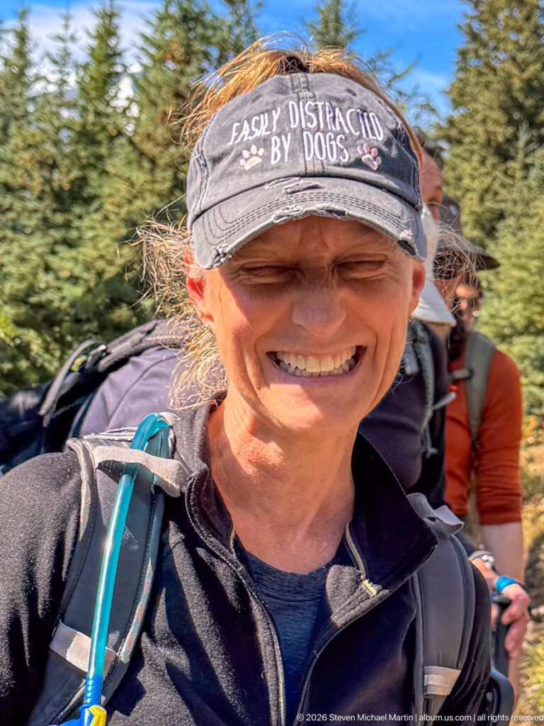 Smiling hiker wearing a visor and backpack on a forested mountain trail, with another hiker behind.