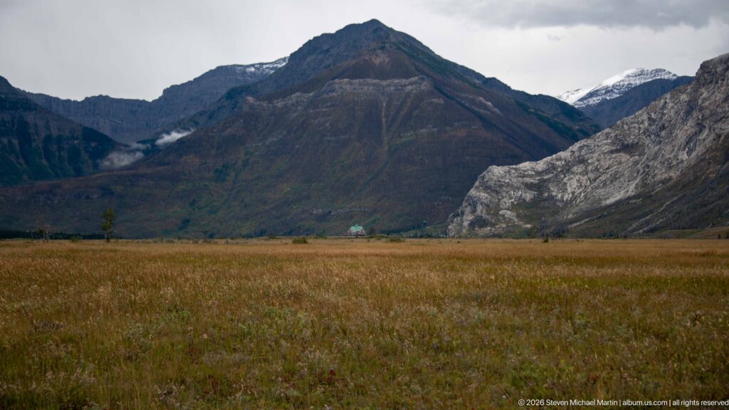 Rust-colored grassland below a dark pyramidal mountain and pale gray rock face, with a tiny building near the mountain base.