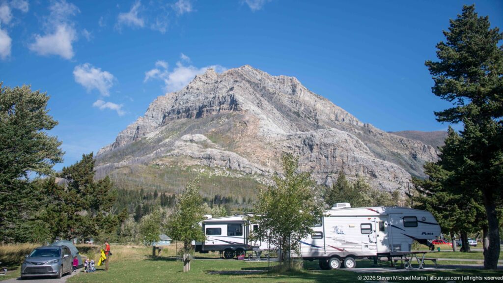 RVs, cars, and campsites on grass beneath a steep layered mountain, with conifer trees framing the Waterton Lakes campground.