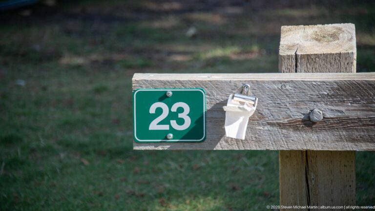 Green campsite marker numbered 23 attached to a weathered wooden rail beside a small white utility fitting.