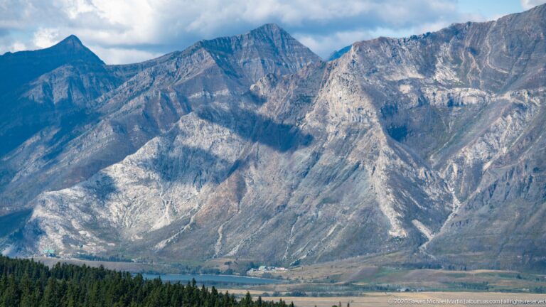Steep gray mountain slopes with exposed layered rock and gullies rising above forest and a narrow developed valley floor.