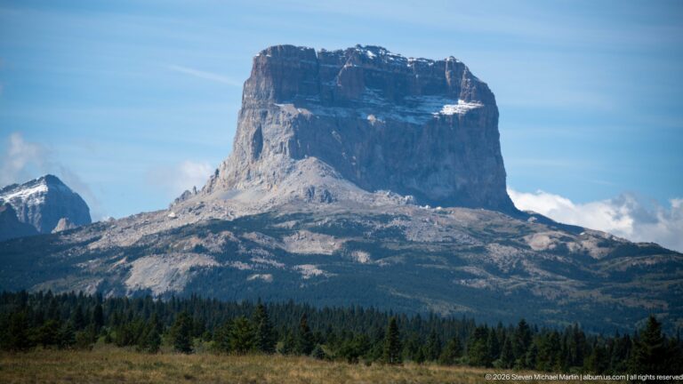 Chief Mountain with a broad flat summit and steep cliffs rising above forest and open foothills under a blue sky.