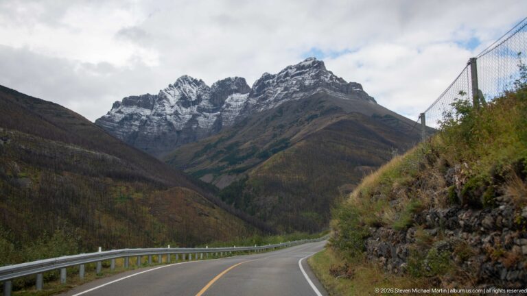 Two-lane mountain road curving toward jagged snow-dusted peaks between steep slopes under broken clouds.