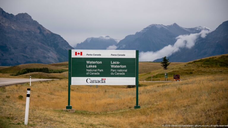Bilingual Waterton Lakes National Park of Canada entrance sign in golden grassland with cloud-wrapped mountains behind.