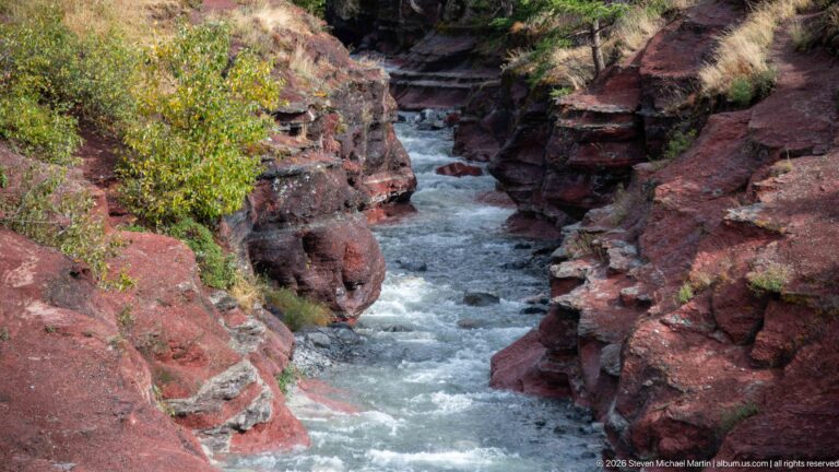 Narrow white-water stream confined between steep dark-red layered rock walls with shrubs along the rim.