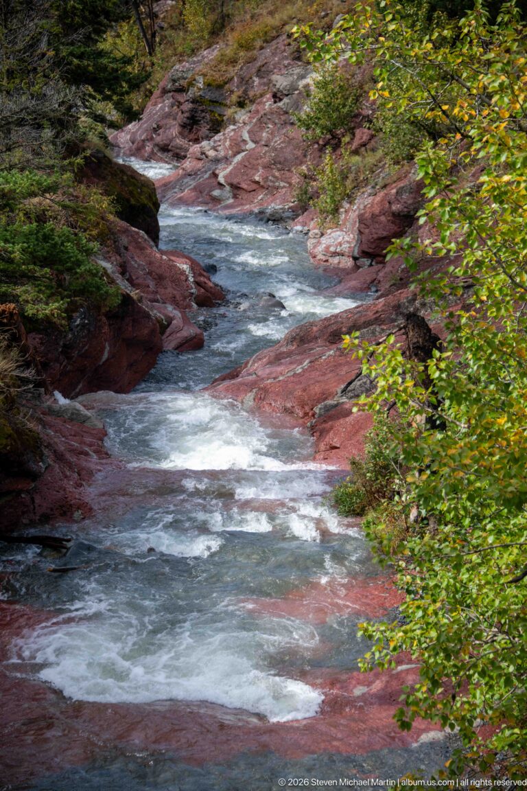 Fast stream with short rapids flowing between red bedrock shelves and green shrubs.