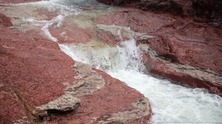 White water dropping through a narrow channel of vivid red and pale layered rock.