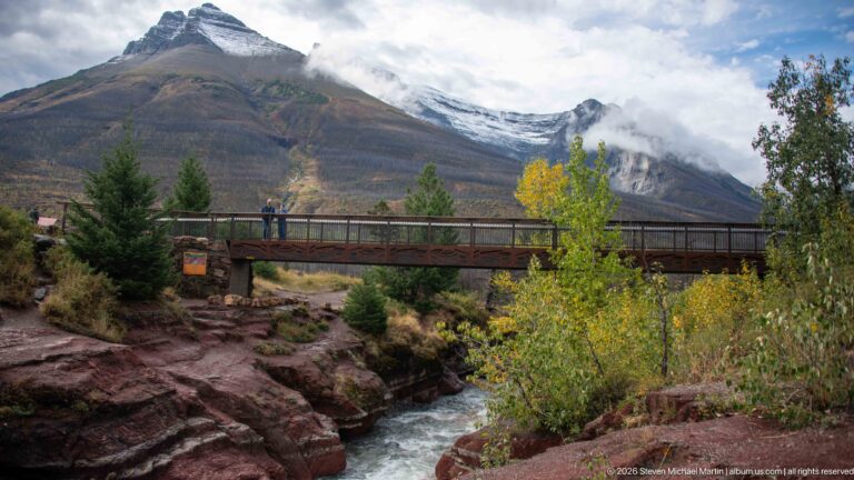 Pedestrian bridge with visitors crossing a fast stream between red rock walls, backed by autumn foliage and cloud-covered mountains.